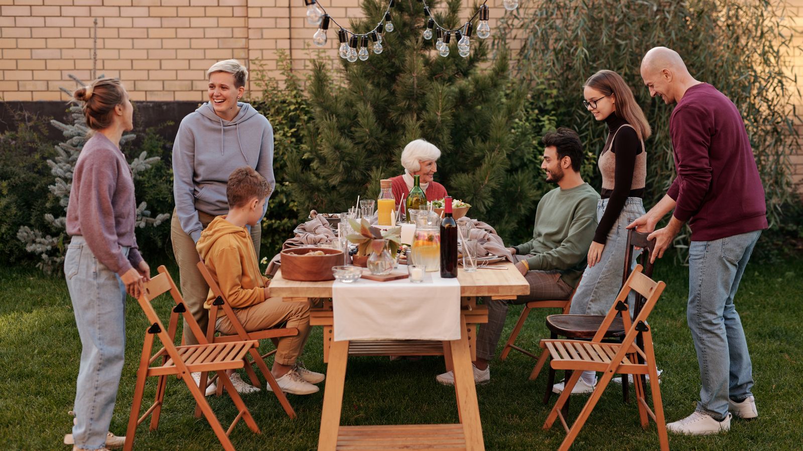Family members talking and eating at a long wooden table in a back garden during a family gathering